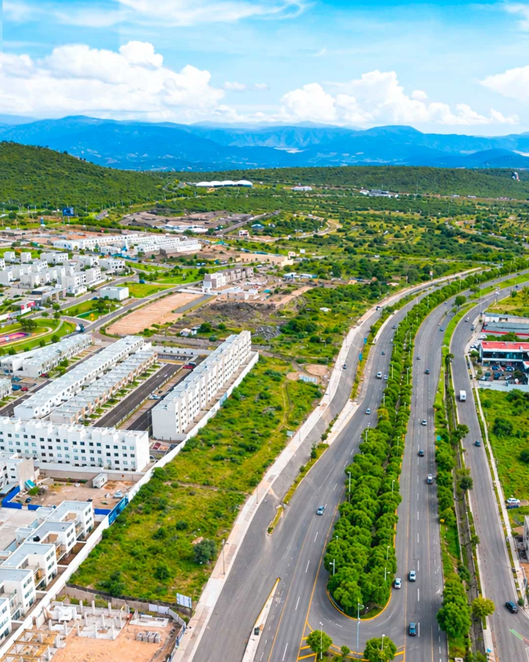 Vista aérea del terreno de Zarú, con una avenida arbolada, construcciones comerciales, desarrollos habitacionales y cerros al fondo, en Nuevo Refugio, Querétaro.